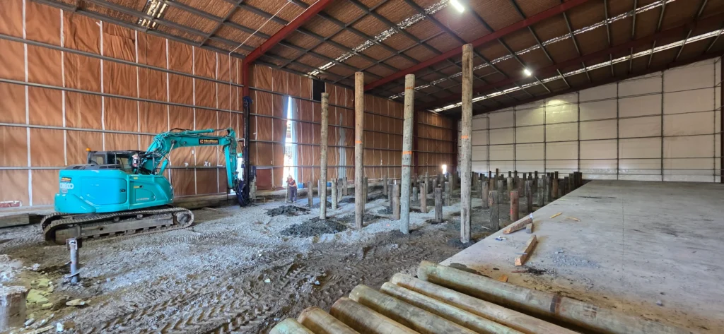 Excavator working inside a warehouse during concrete strengthening, with timber piles in place